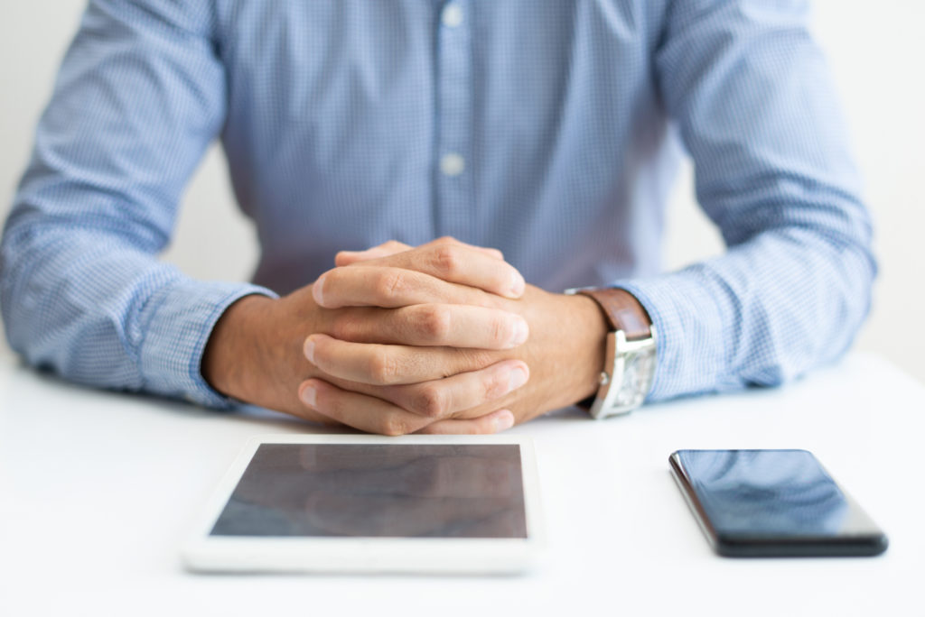 Closeup of man sitting at desk with tablet and smartphone. Business man sitting with his hands clasped. Technology and communication concept. Cropped front view.